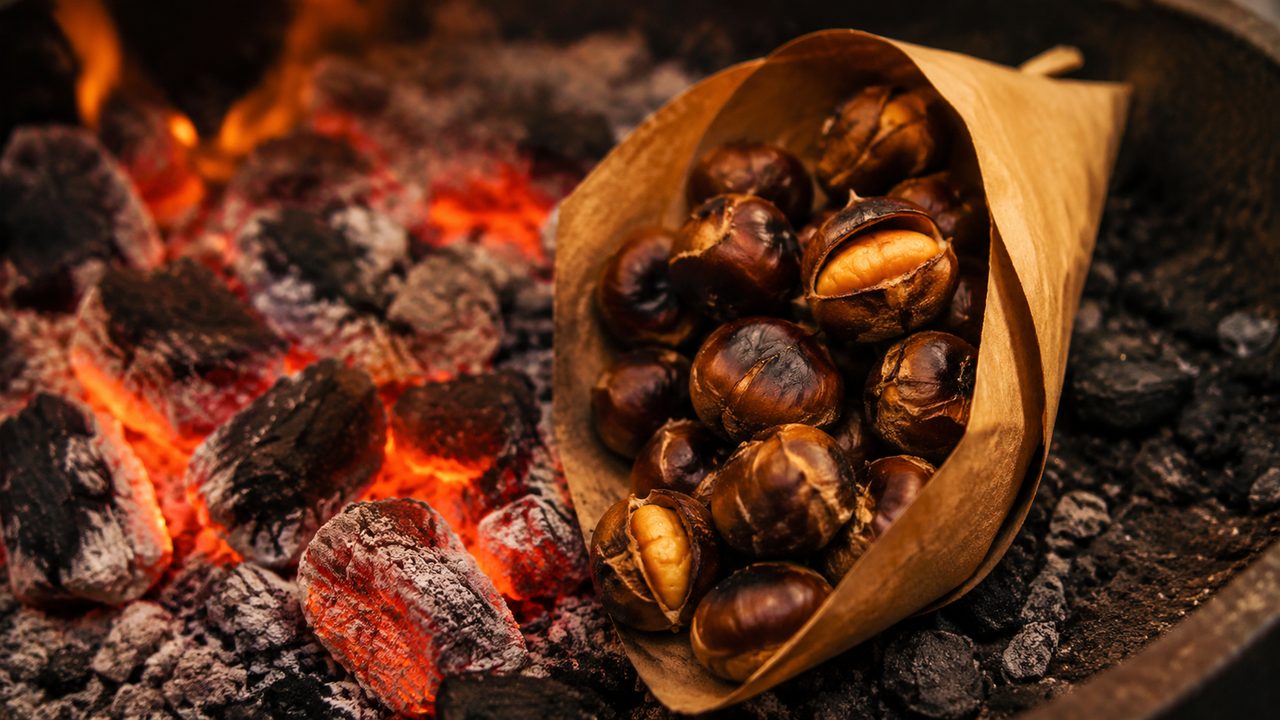 A street vendor's pan of scored chestnuts roasting over coals, a paper cone of them beside it